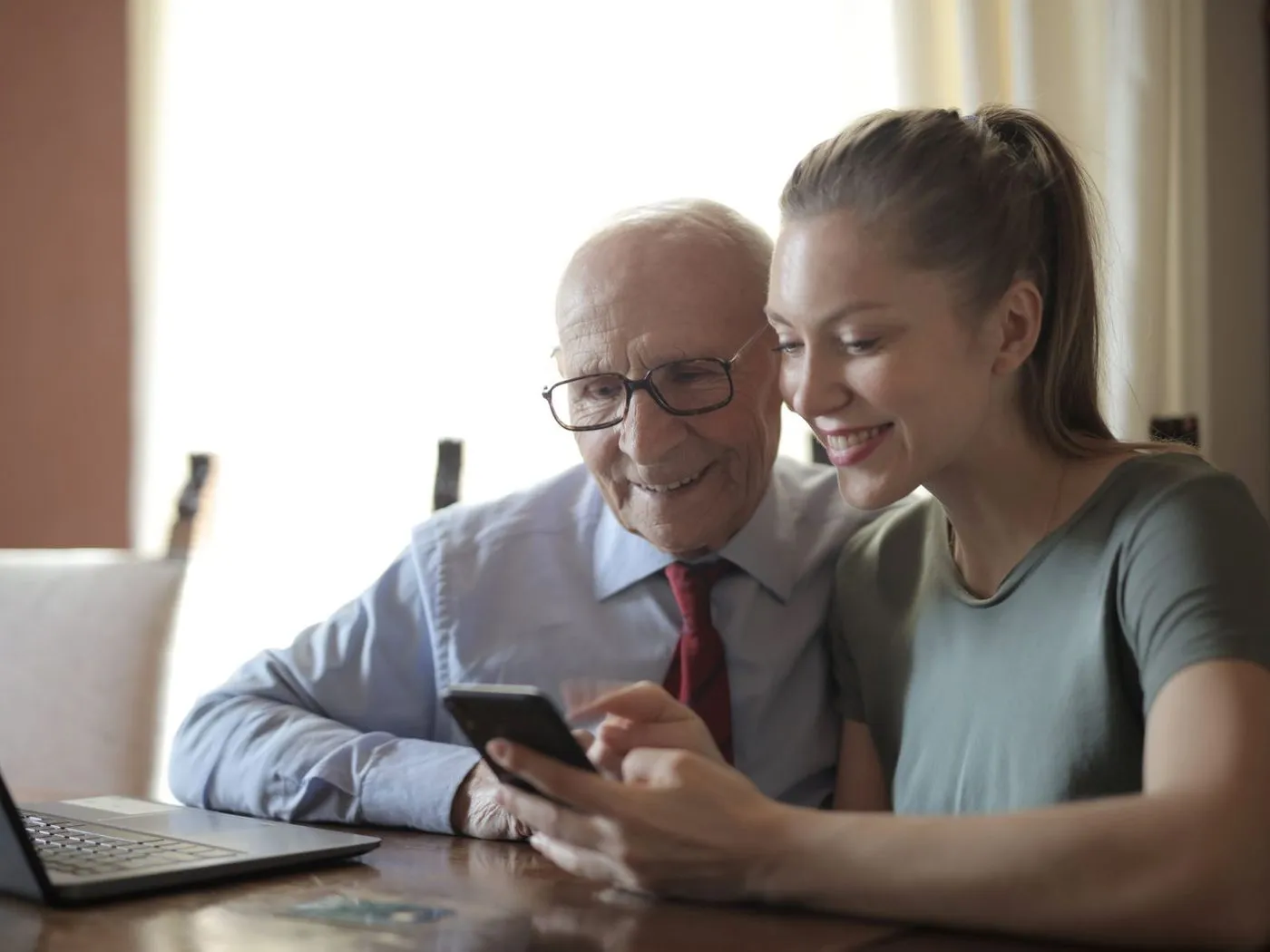 Een jongere vrouw laat een oudere man iets zien op een telefoon, met een laptop op tafel.